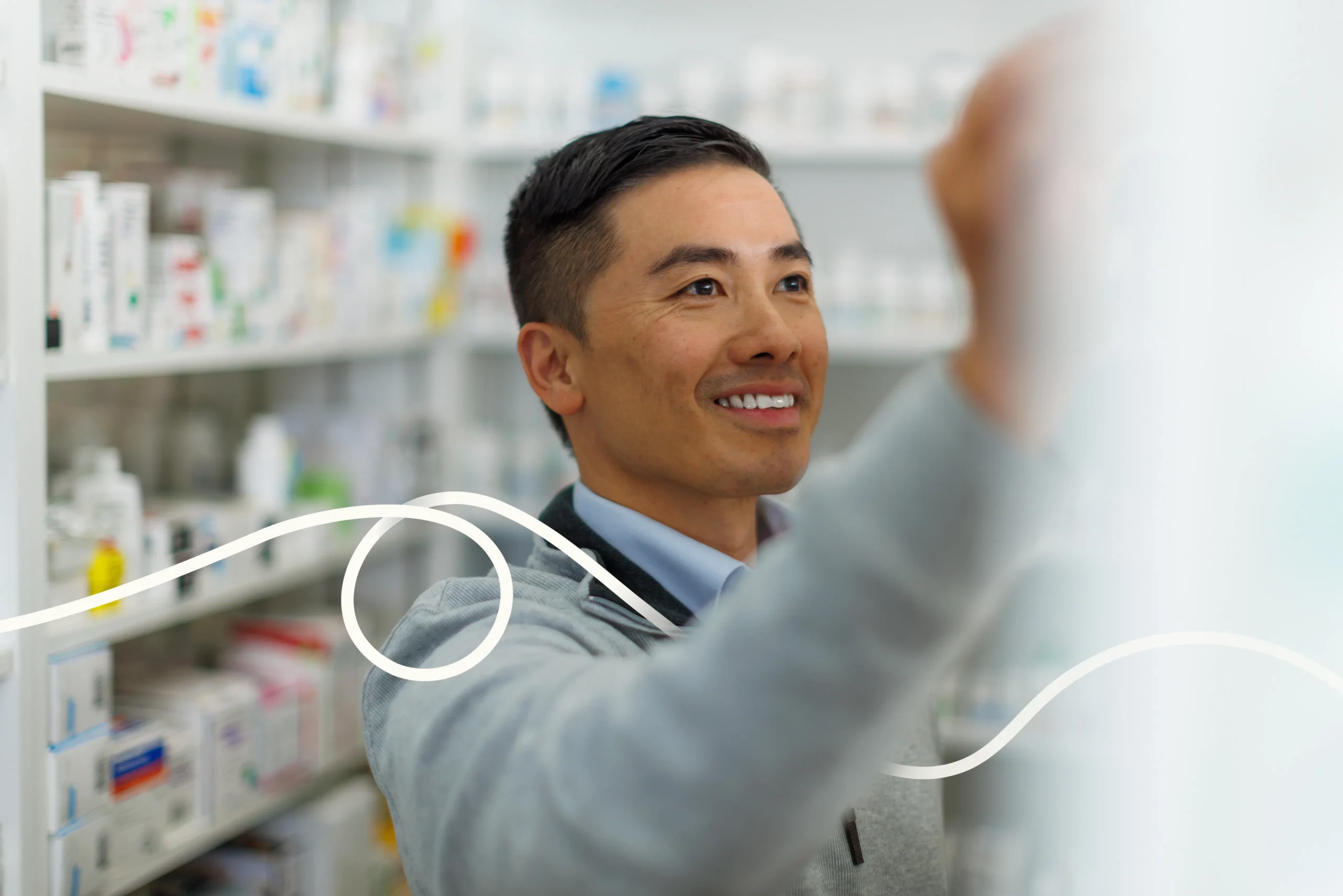 A smiling Asian man wearing a grey sweater reaches up to select a product from a stocked pharmacy shelf, with a decorative white wavy line overlay in the foreground.