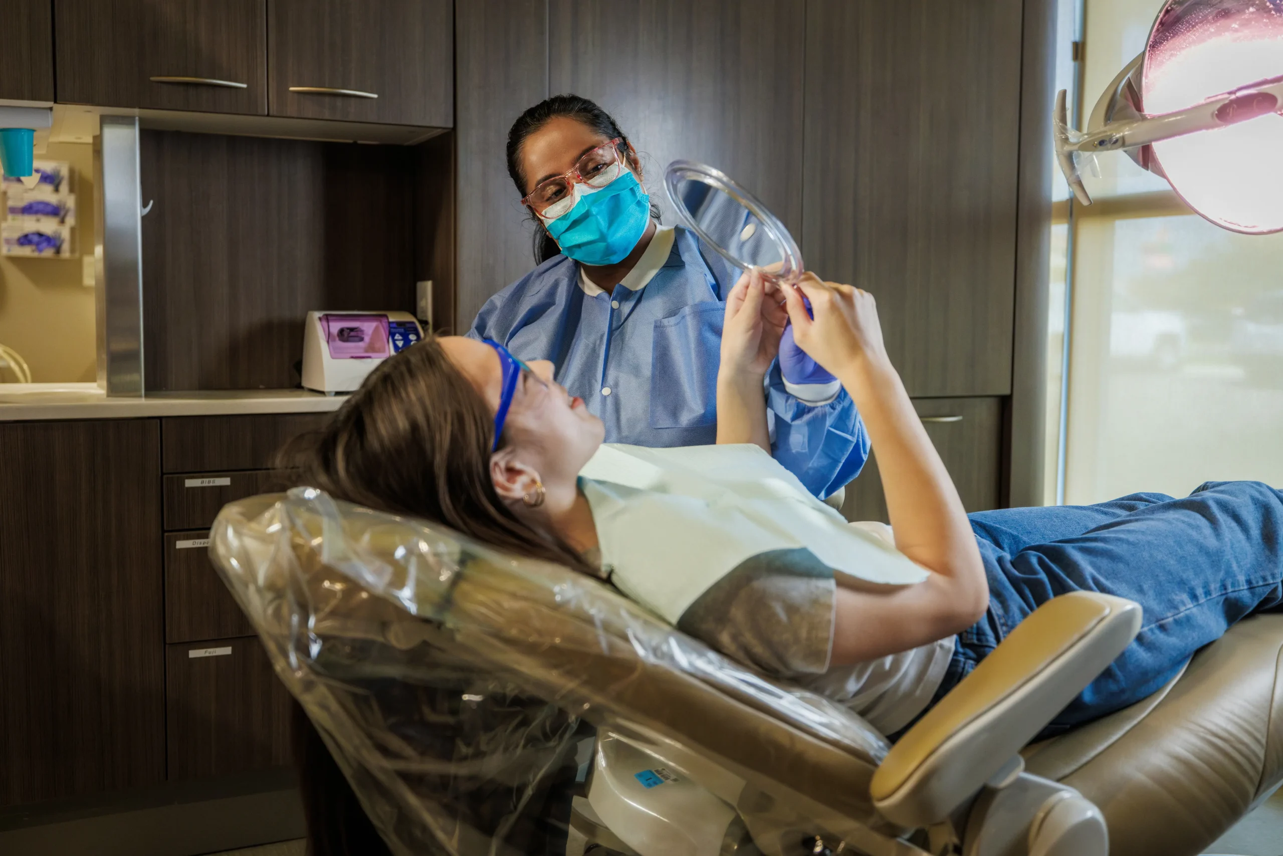 A woman wearing a hijab writes in a notebook in the foreground while a blurred man gestures on a sofa in the background.