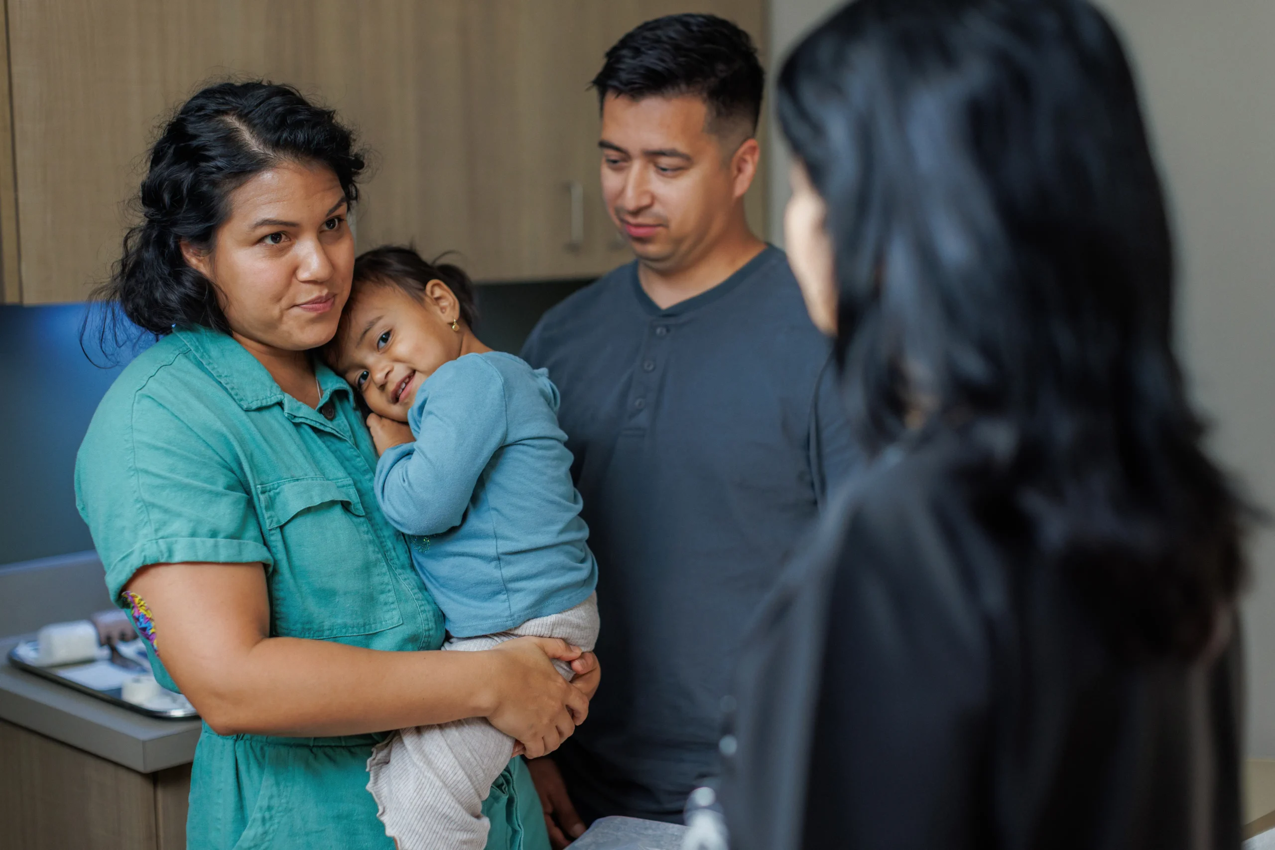 A mother holding her young daughter stands next to a man in a medical exam room, listening to a female healthcare provider whose back is turned; the mother has a colorful bandage on her arm.