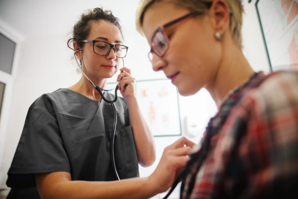 A female doctor in dark grey scrubs uses a stethoscope to examine a female patient wearing a plaid shirt.